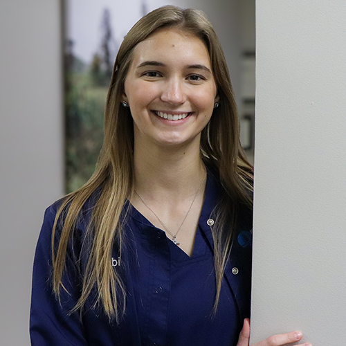 Dental Assistant Abbi leaning against a hallway wall in blue scrubs
